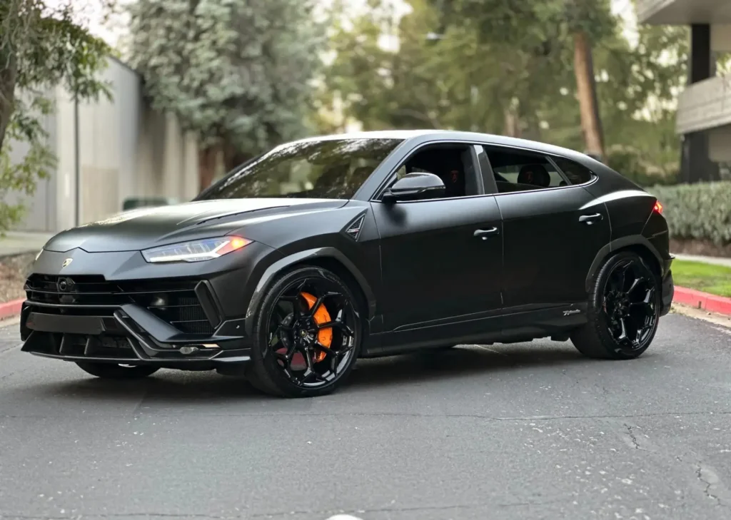 Matte black Lamborghini Urus luxury SUV parked on a quiet tree-lined street, featuring black alloy wheels, orange brake calipers, and a sleek performance-focused design.