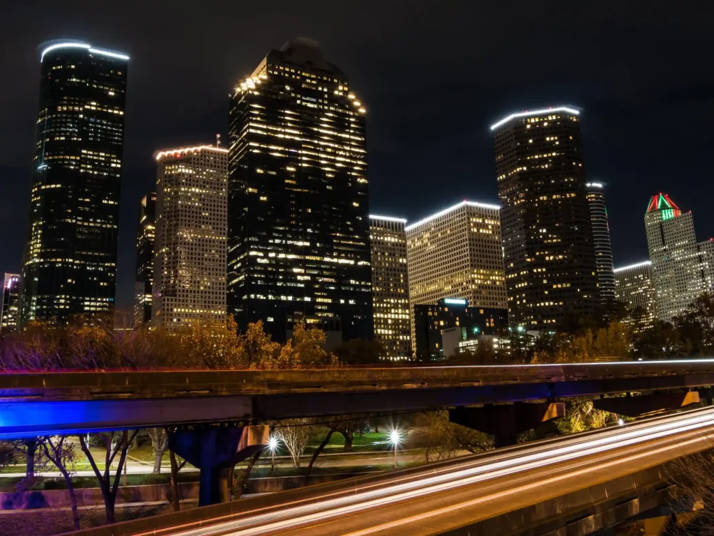 Night view of the Houston skyline with illuminated skyscrapers and long-exposure highway light trails in downtown Houston.